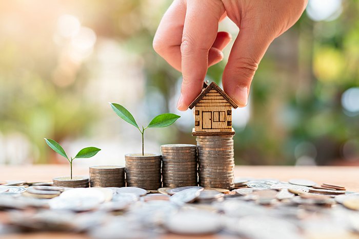 Hand placing a small wooden house on stacked coins in California, surrounded by coins and sprouting plants, symbolizing growth and investment.