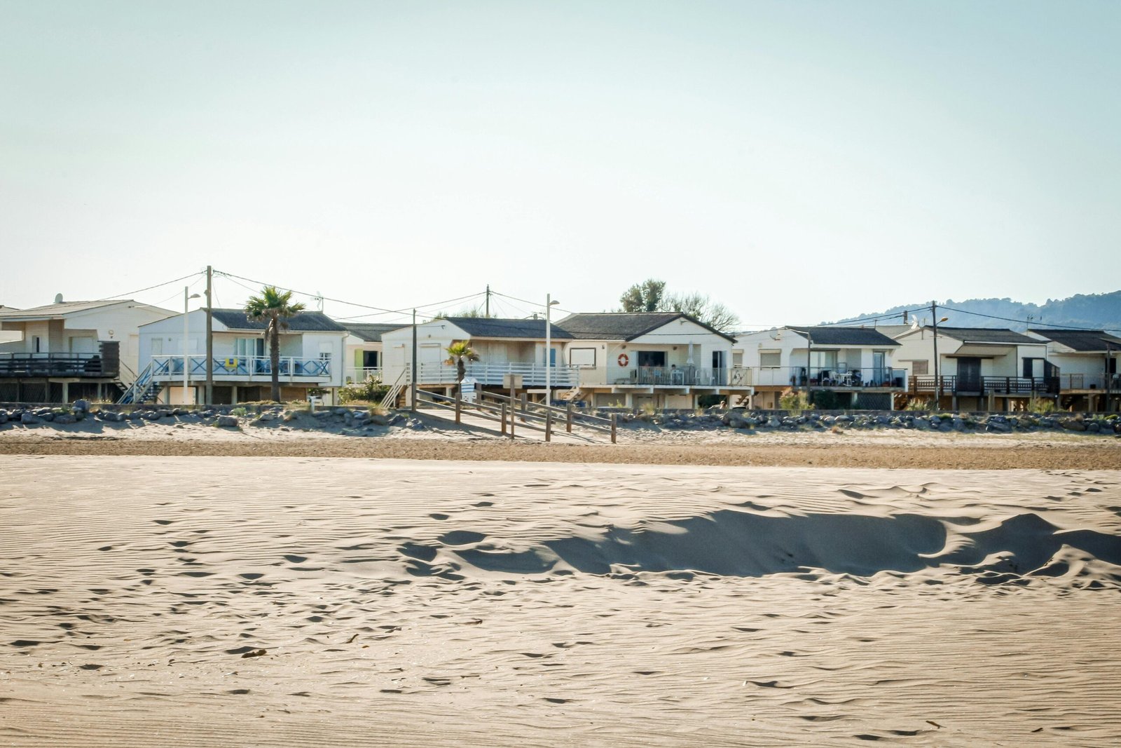 Beachfront view in California of white cabins on stilts with sand, blue sky, and distant hills, creating a serene coastal vibe.