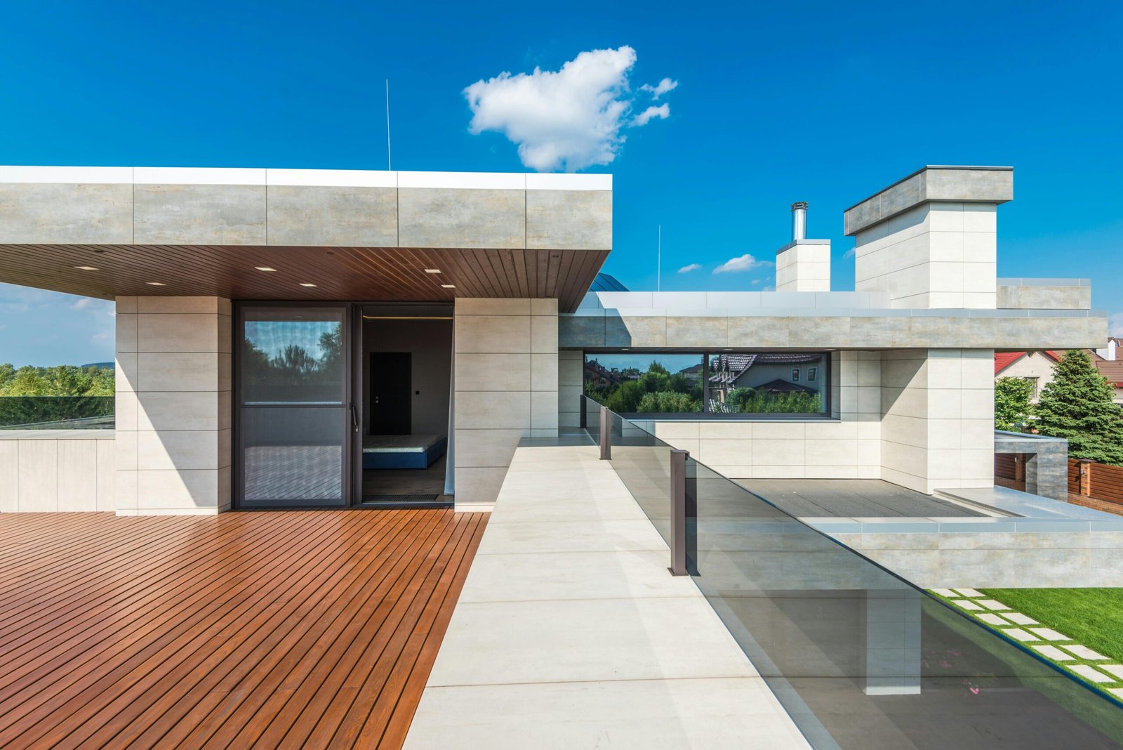 Modern Southern California rooftop terrace with wood floors, beige stone walls, glass railings, and plants under a clear blue sky.
