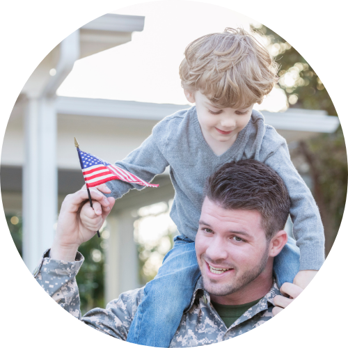 A smiling man in military uniform carries a young boy on his shoulders, with the boy holding a small American flag, showing pride and happiness.