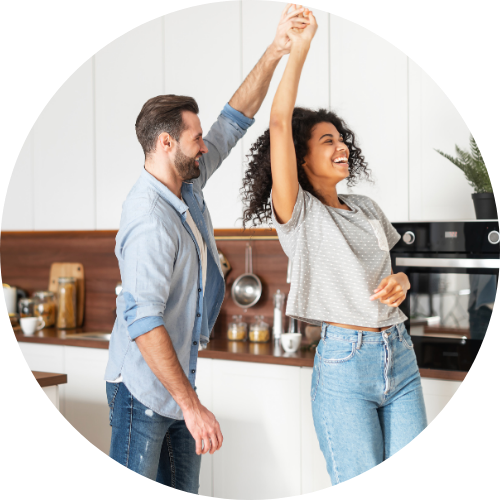 A couple dances happily in a modern kitchen, with the man twirling the woman as sunlight fills the cozy, well-equipped space.