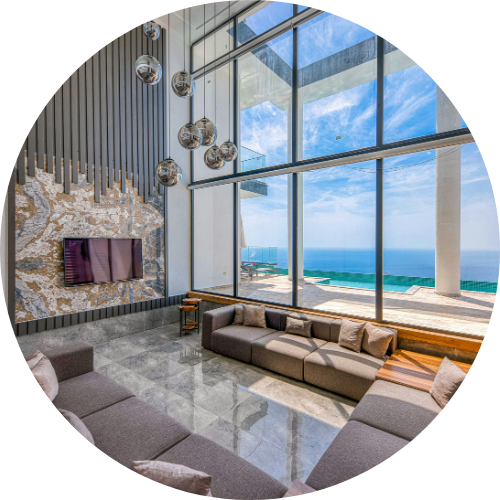 Modern living room with floor-to-ceiling windows showing ocean views, gray sofas, marble floors, and a wall with a TV.