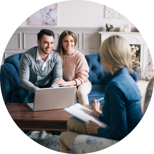 A couple sits on a blue sofa, smiling and talking to a woman with a notepad, with a laptop on the table in a warm, friendly setting.