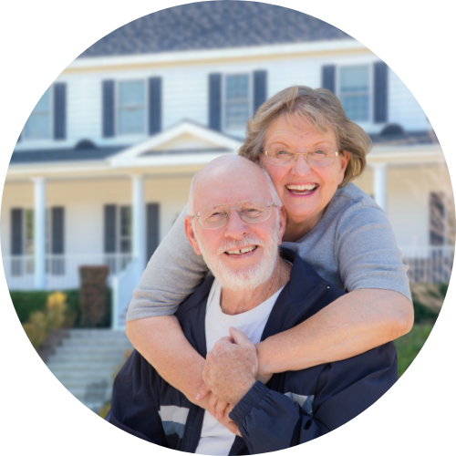 An elderly couple smiles warmly in front of a large white house, with the woman hugging the man from behind,